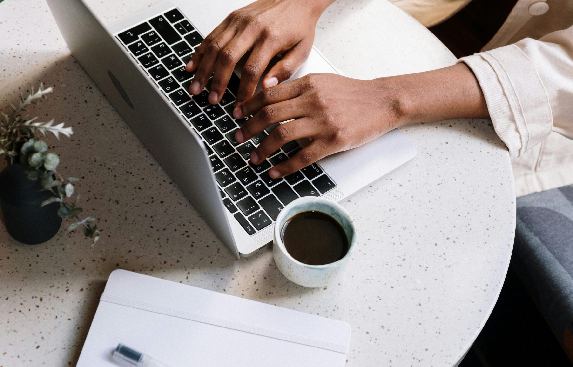 A person typing on a laptop at a café table with coffee and a notebook.